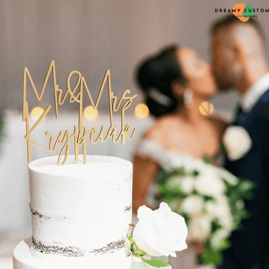 A beautifully personalized gold cake topper crowns a simple yet elegant wedding cake, while the newlyweds share a romantic kiss in the background. A perfect touch for a memorable wedding celebration.