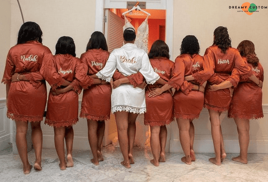 The bride and her bridesmaids stand arm-in-arm in personalized satin robes, facing her wedding dress. A heartfelt pre-wedding moment filled with love, unity, and anticipation for the big day.
