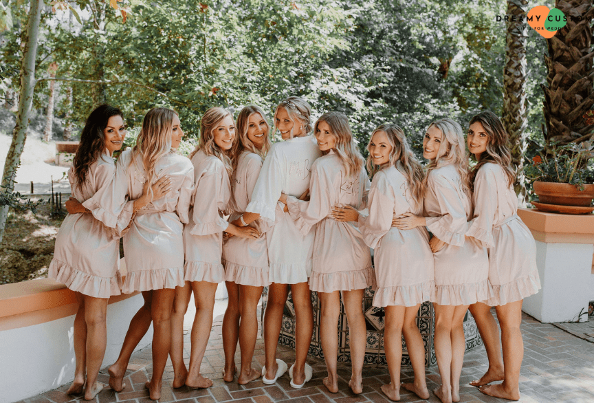 A joyful bridal party poses in matching pink robes with the bride in white, capturing a beautiful and coordinated pre-wedding moment.