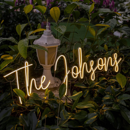 A warm, glowing personalized neon sign reading "The Johnsons" nestled among lush greenery, creating a romantic and modern wedding ambiance. A perfect touch for outdoor celebrations.