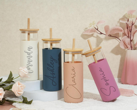 The image features four custom glass tumblers with bamboo lids and straws. Each tumbler is personalized with a different name: Amanda, Ashley, Clara, and Sophia, showcasing a variety of pastel colors.