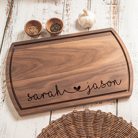 A personalized walnut wood cutting board engraved with "sarah ♥ jason" displayed on a rustic wooden table, with small bowls of spices and a garlic clove in the background, creating a cozy, kitchen setting.