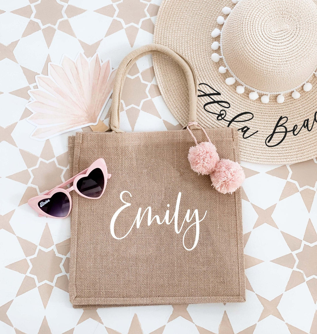 The image shows a personalized burlap tote bag with the name "Emily" elegantly written in white, adorned with pink pom-poms. The bag is surrounded by a summer hat, sunglasses, and a fan, giving it a stylish, vacation-ready vibe.
