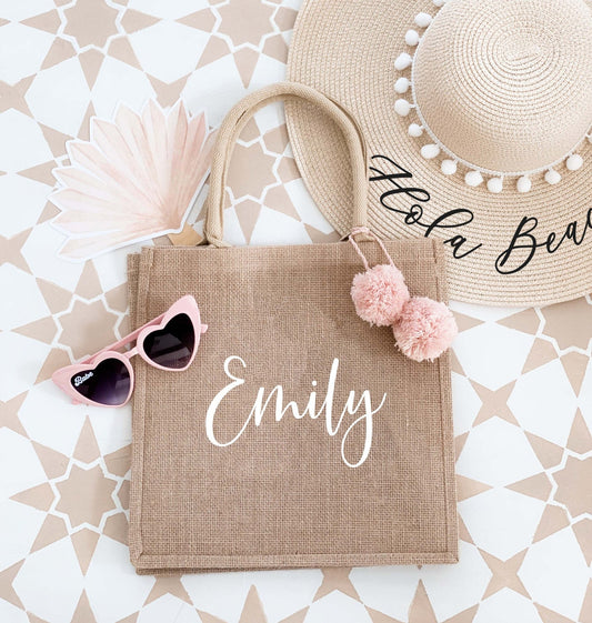 The image shows a personalized burlap tote bag with the name "Emily" elegantly written in white, adorned with pink pom-poms. The bag is surrounded by a summer hat, sunglasses, and a fan, giving it a stylish, vacation-ready vibe.
