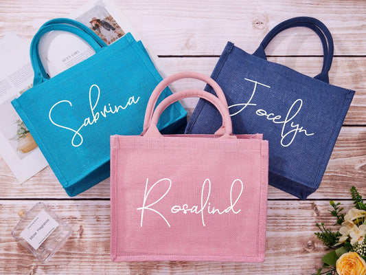 The image displays three personalized burlap tote bags in different colors: teal, pink, and navy. Each bag features a name written in elegant white script, perfect for gifting or personal use.