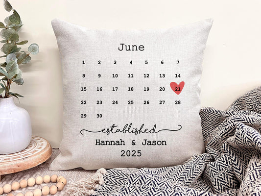 A personalized wedding pillow with a June calendar, highlighting the 21st with a red heart. Below are the names "Hannah & Jason" and "2025." The pillow rests on a cozy surface with a textured look.