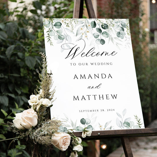 The image displays a beautiful wedding welcome sign for Amanda and Matthew, featuring elegant greenery and soft florals. The sign is displayed on an easel, adding a charming touch to the outdoor wedding setting.