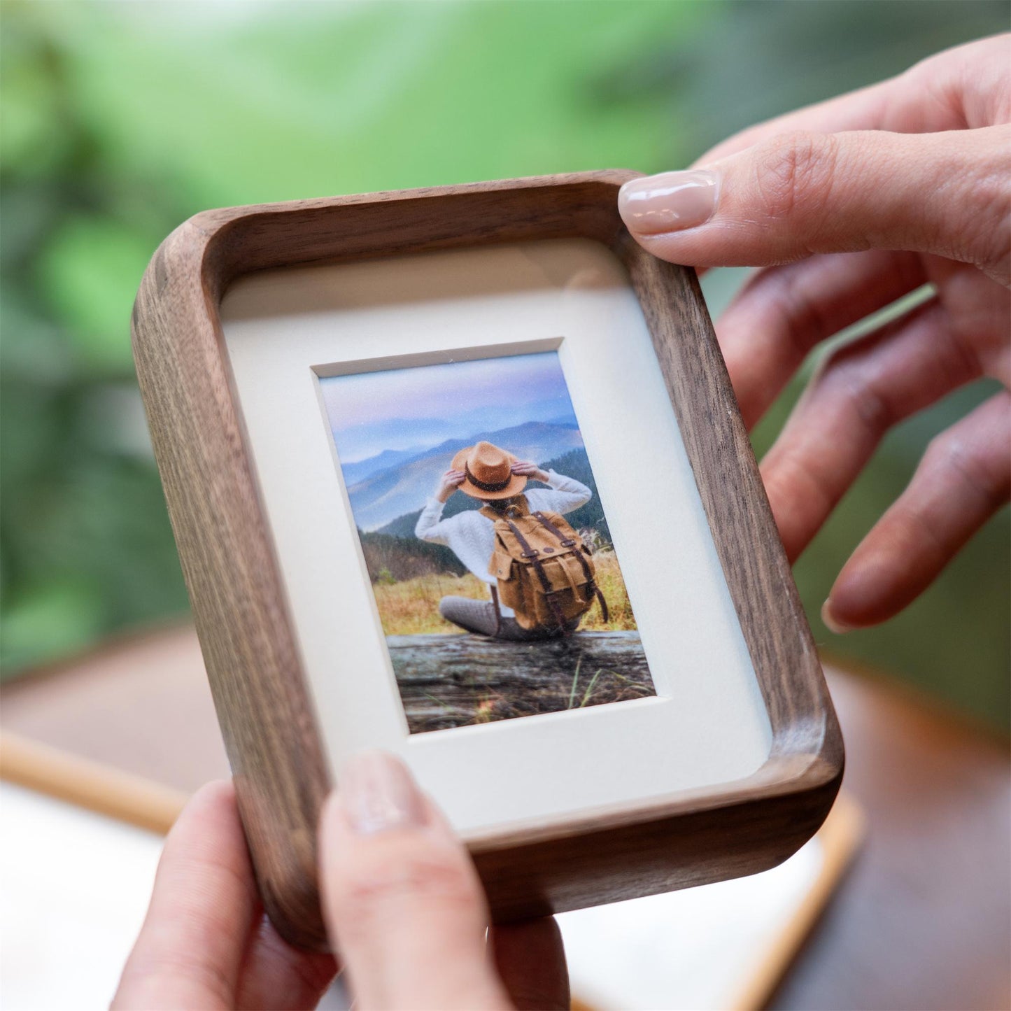 A walnut wood mini photo frame holding a photo of a person sitting outdoors, looking at mountains. The frame features a clean, modern design with a white matting around the photo.
