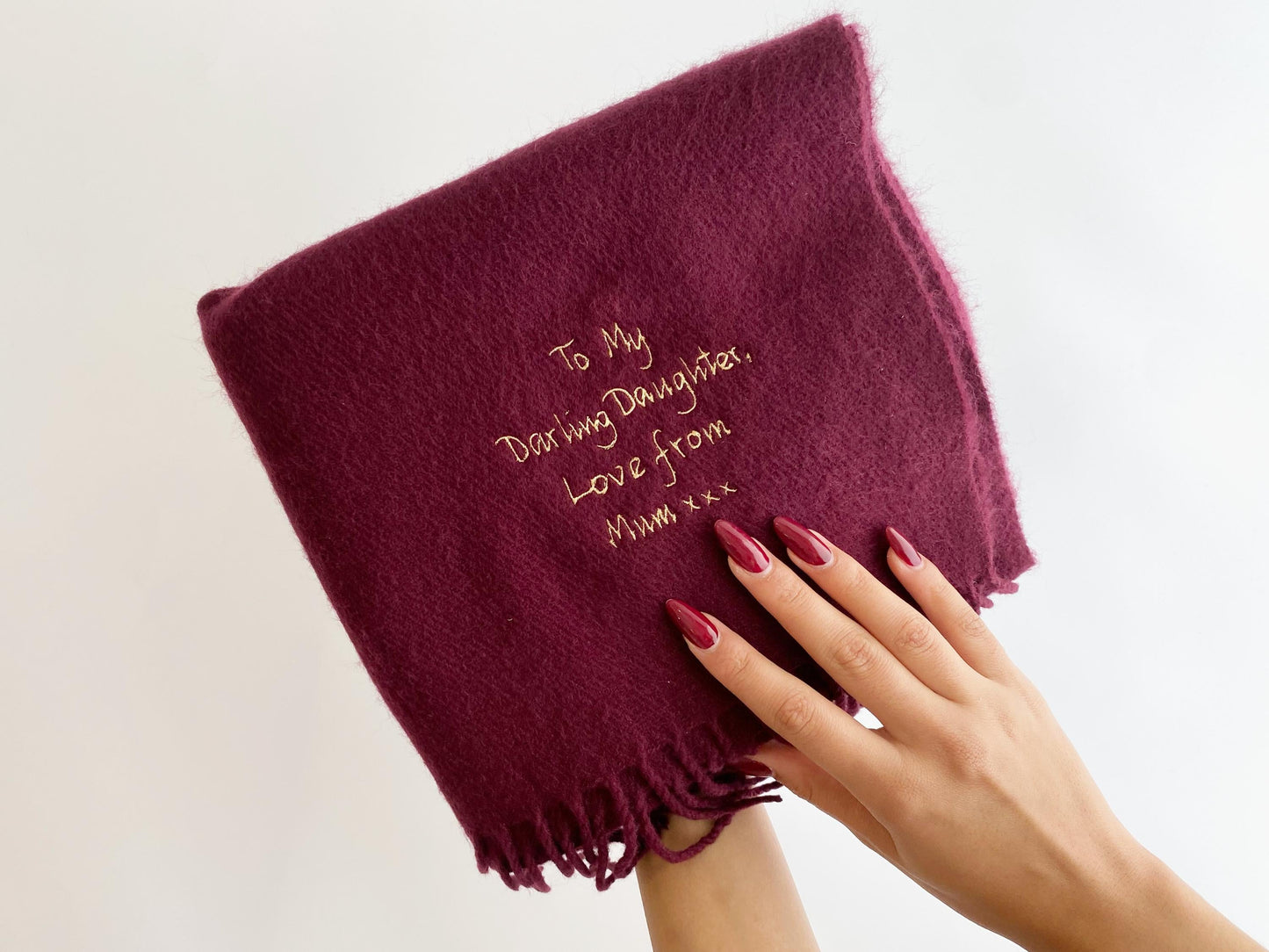 A hand with red nail polish holds a red personalized scarf embroidered with "To My Darling Daughter, Love from Mum xxx" against a white background.