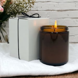 A lit amber jar personalized wedding candle next to a white gift box with a black ribbon, set against a white brick wall with a bouquet of flowers in the background.