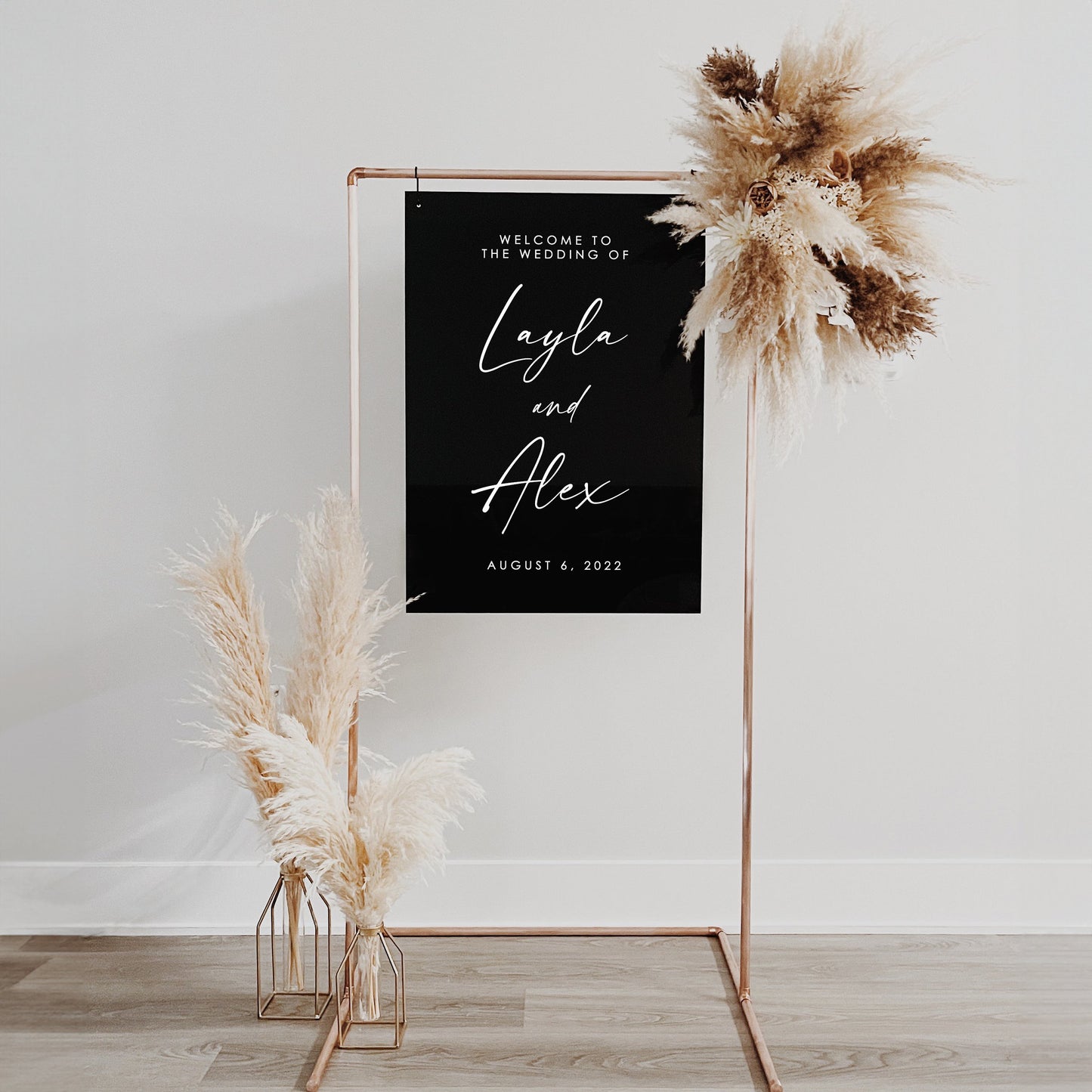 Elegant black wedding welcome sign on a copper frame, featuring "Layla and Alex" with pampas grass decoration.