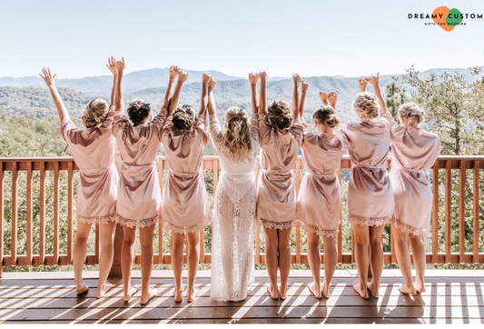 Bridesmaids in matching satin robes and a bride in a white lace robe raise their hands joyfully against a scenic mountain backdrop. A perfect bridal party moment, exuding elegance and excitement for the big day.