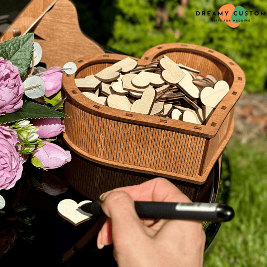 Heart-shaped wooden guest book box filled with blank wooden hearts, with a guest writing a message on one heart. A unique and interactive wedding guest book alternative for capturing heartfelt wishes.