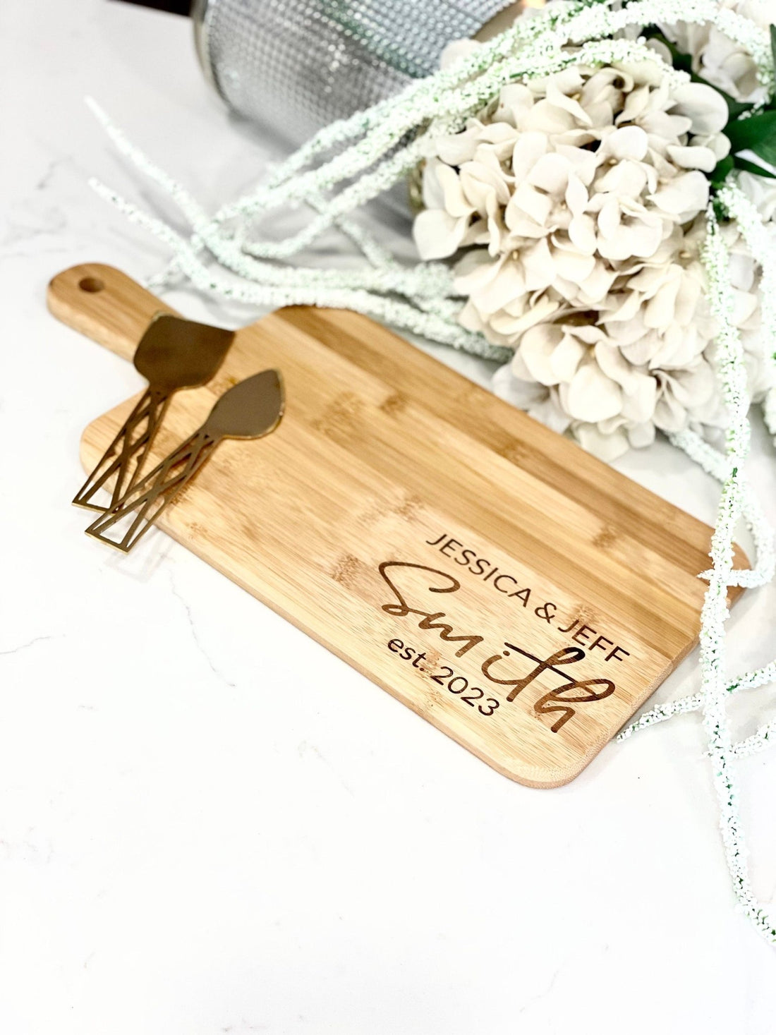 A personalized bamboo charcuterie board engraved with "JESSICA & JEFF SMITH est. 2023" alongside two gold cheese knives, set against a backdrop of white hydrangea flowers, reflecting an elegant and modern look for spring gatherings.