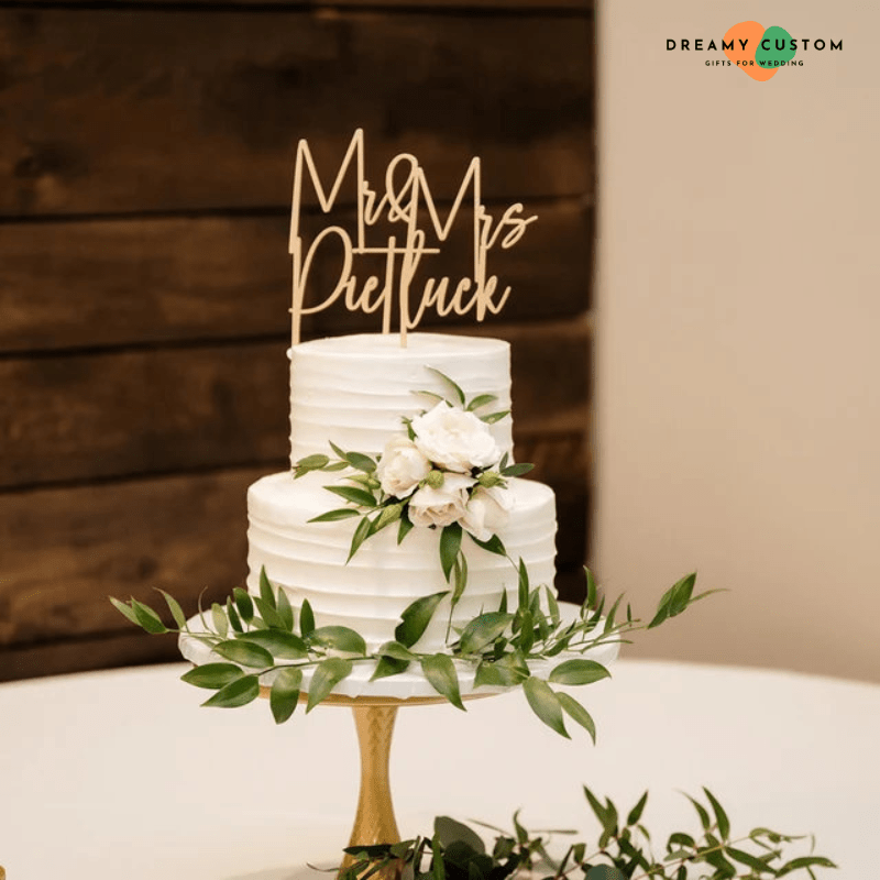 Wedding cake with a "Mr & Mrs Pietluck" wooden cake topper, adorned with white flowers and green leaves, placed on a tiered white cake.