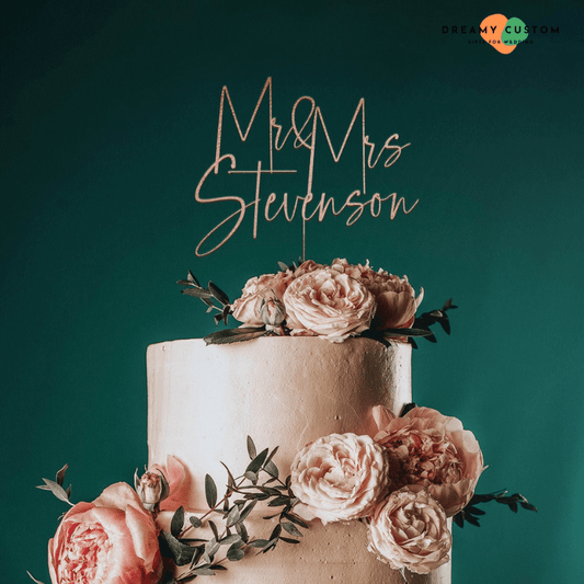 Personalized wedding cake topper with the names 'Mr & Mrs Stevenson' in rose gold, placed on a two-tier wedding cake decorated with blush pink peonies and greenery.