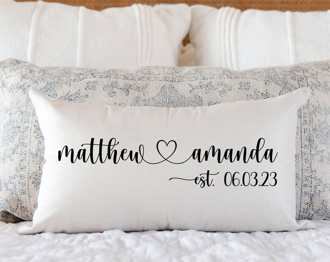 A personalized wedding pillow with the names "Matthew" and "Amanda" separated by a heart symbol, and "est. 06.03.23" written in elegant script, placed against a soft, neutral-colored bedspread.