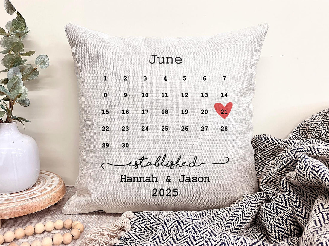 A personalized wedding pillow with a June calendar, highlighting the 21st with a red heart. Below are the names "Hannah & Jason" and "2025." The pillow rests on a cozy surface with a textured look.