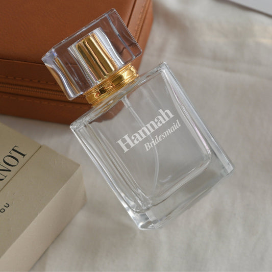 A personalized perfume bottle engraved with "Hannah Bridesmaid" on a glass bottle, placed next to a book and a brown leather bag on a white fabric background.