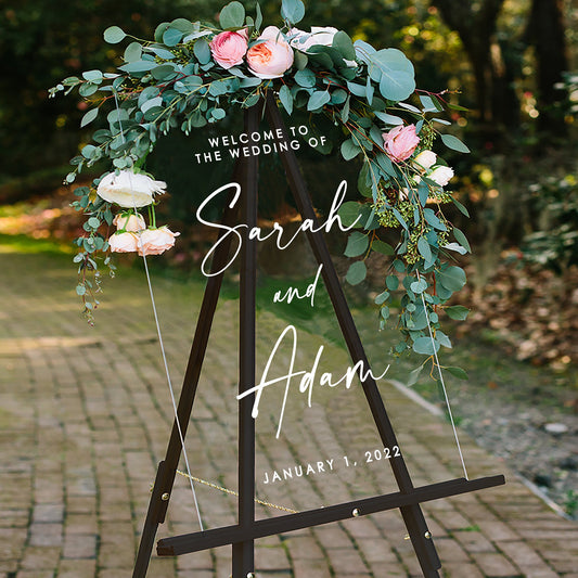 Custom wedding welcome sign with elegant floral decoration and "Sarah and Adam" text on clear acrylic board.